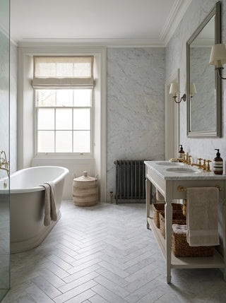 A bright, modern bathroom featuring a white freestanding bath, a white double-sink vanity with gold fixtures, white marble tiles on the walls and floor and a large wall mirror.