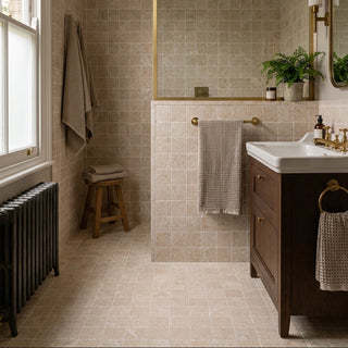A styled bathroom interior with beige marble tiled walls and floor, a dark wood vanity with a white sink, brass fixtures, a small wooden stool and towels hanging on a towel bar and wall.