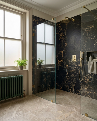 A modern bathroom shower area with dark black marble effect walls and floor, framed by clear glass panels. A gold shower head and controls are mounted on the wall, with a folded towel hanging on a small shelf.