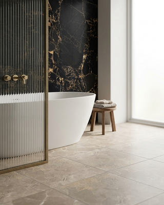 A modern bathroom interior with a freestanding white bathtub, a ribbed glass shower screen with brass hardware, a black marble wall with gold veining, and light stone floor tiles. A small wooden stool with folded towels sits beside the tub.
