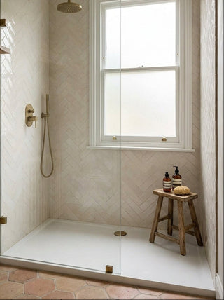 A modern walk-in shower enclosure with a frameless glass door, featuring a white tiled shower base, a herringbone-patterned beige tile wall, a brass shower head and controls, and a small wooden stool holding toiletries next to a white-framed window.
