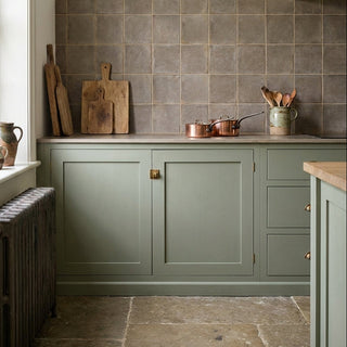 A rustic kitchen corner with sage green cabinets, an olive tiled backsplash, and copper cookware on a stone countertop.